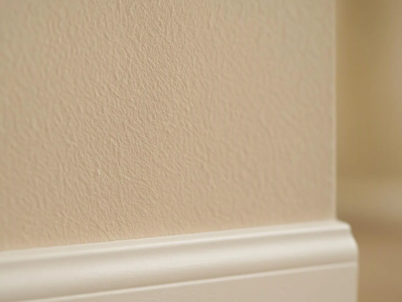 Close-up of a beige textured wall with white baseboard trim along the bottom