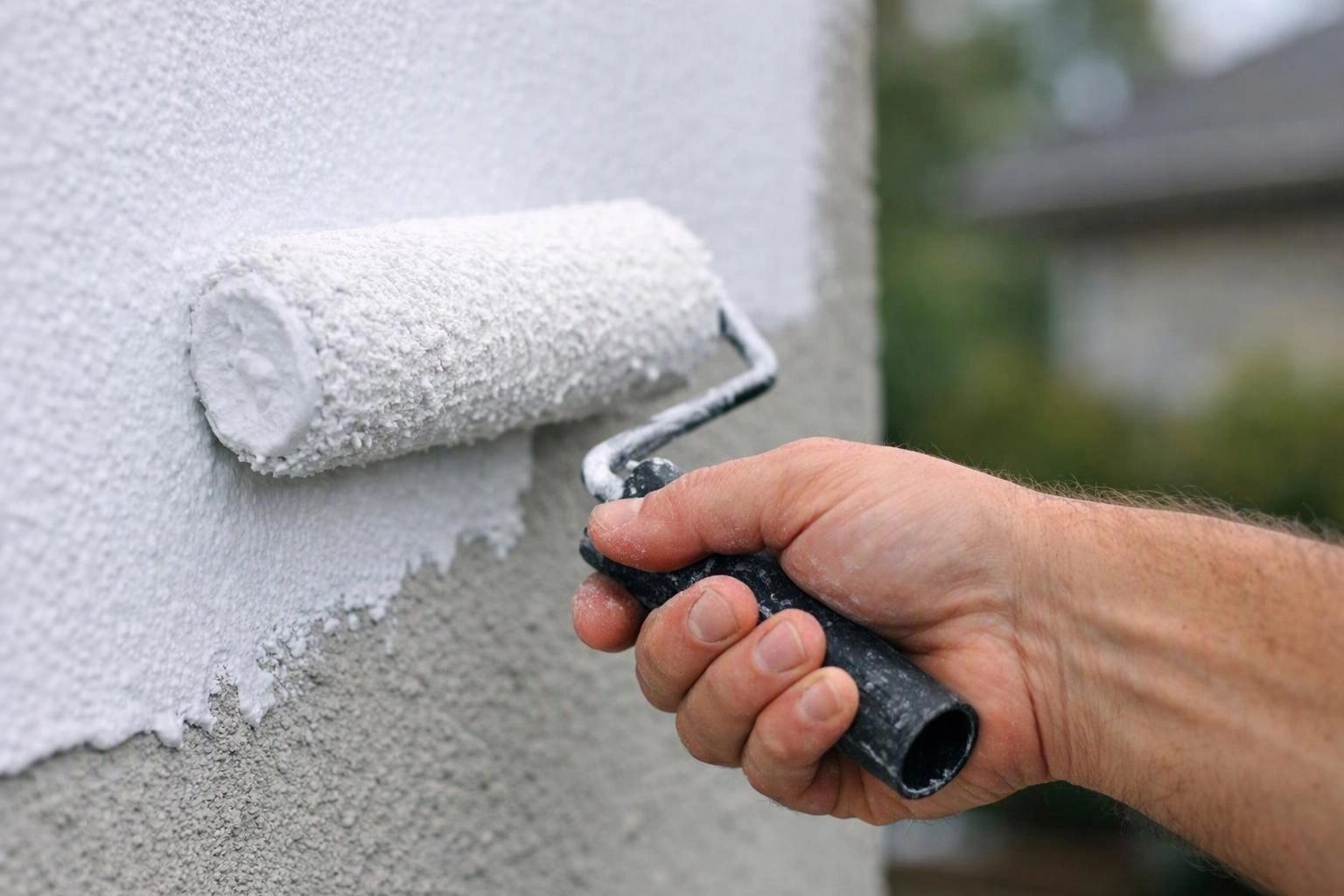 A hand rolls white paint onto a textured exterior wall with a paint roller