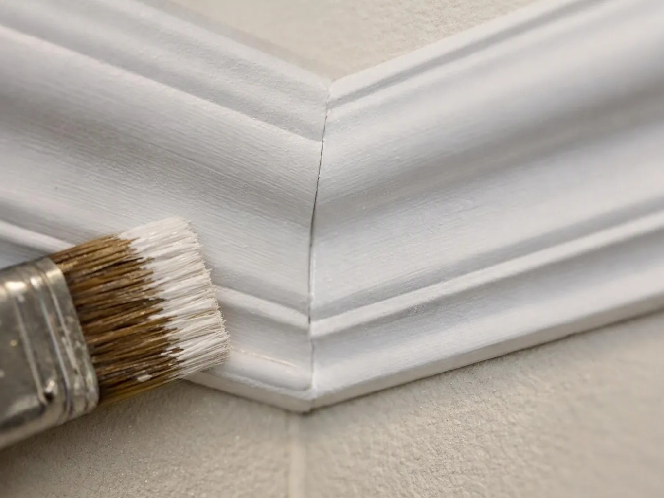 A paintbrush applies white paint to a corner of decorative wall molding