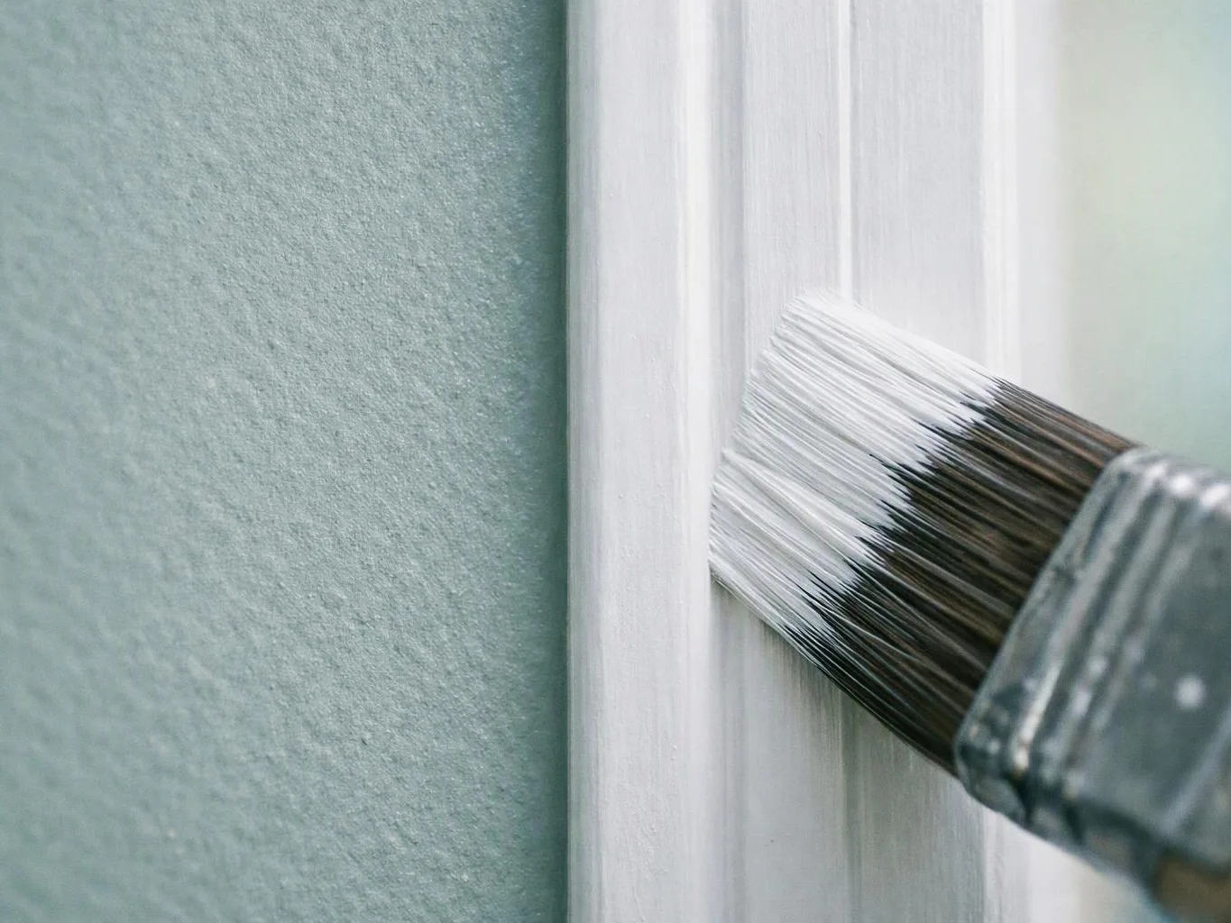A paintbrush applies white paint to a door frame beside a textured green wall