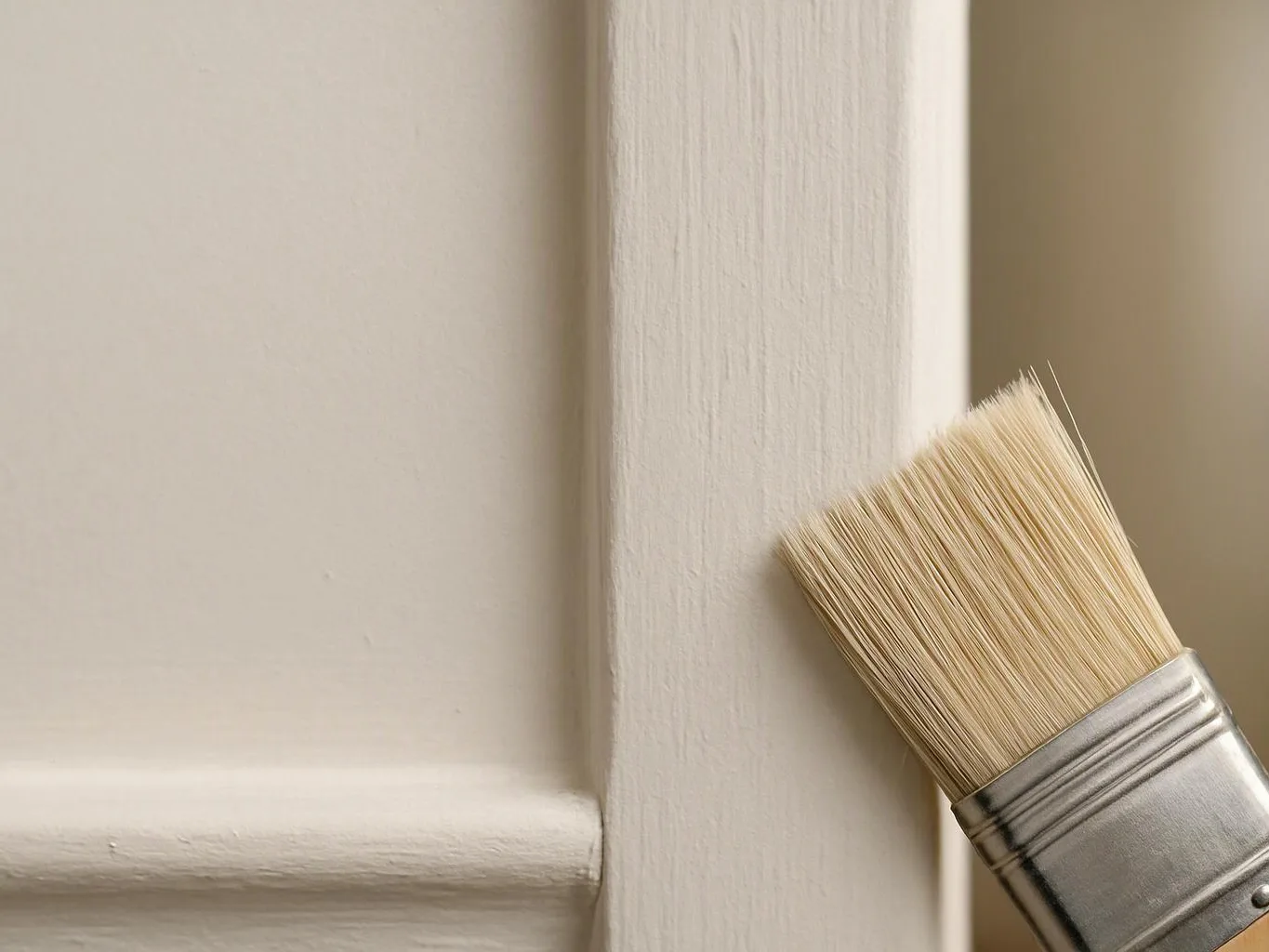 A paintbrush touches a beige wooden wall trim in a close-up interior scene